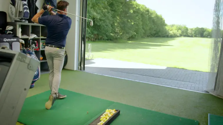 Golflehrer beim Abschlag in der Trainerbox, durch das offene Tor der Blick auf die Driving Range