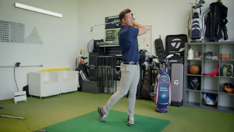 Golflehrer im Durchschwung in der Trainerbox, im Hintergrund Golfbags, Schläger und Trainingsgeräte